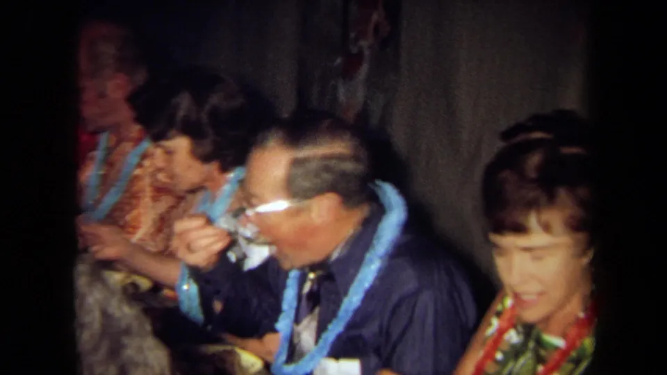 Adults in leis and patterned shirts eat and converse at a crowded indoor dinner table in archival 1971 California home-movie footage.