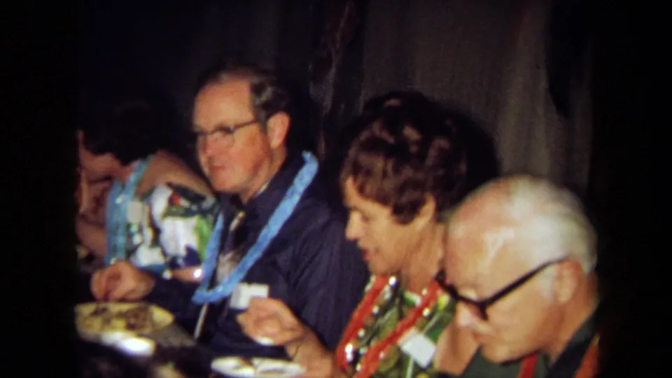 A row of adult dinner guests, some wearing leis and eyeglasses, sit closely at a dim table in 1971 California 8mm footage.