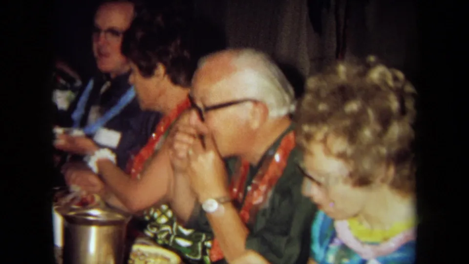 Older adults and nearby guests in leis sit at a crowded table with food and tableware in a dim 1971 California home-movie scene.