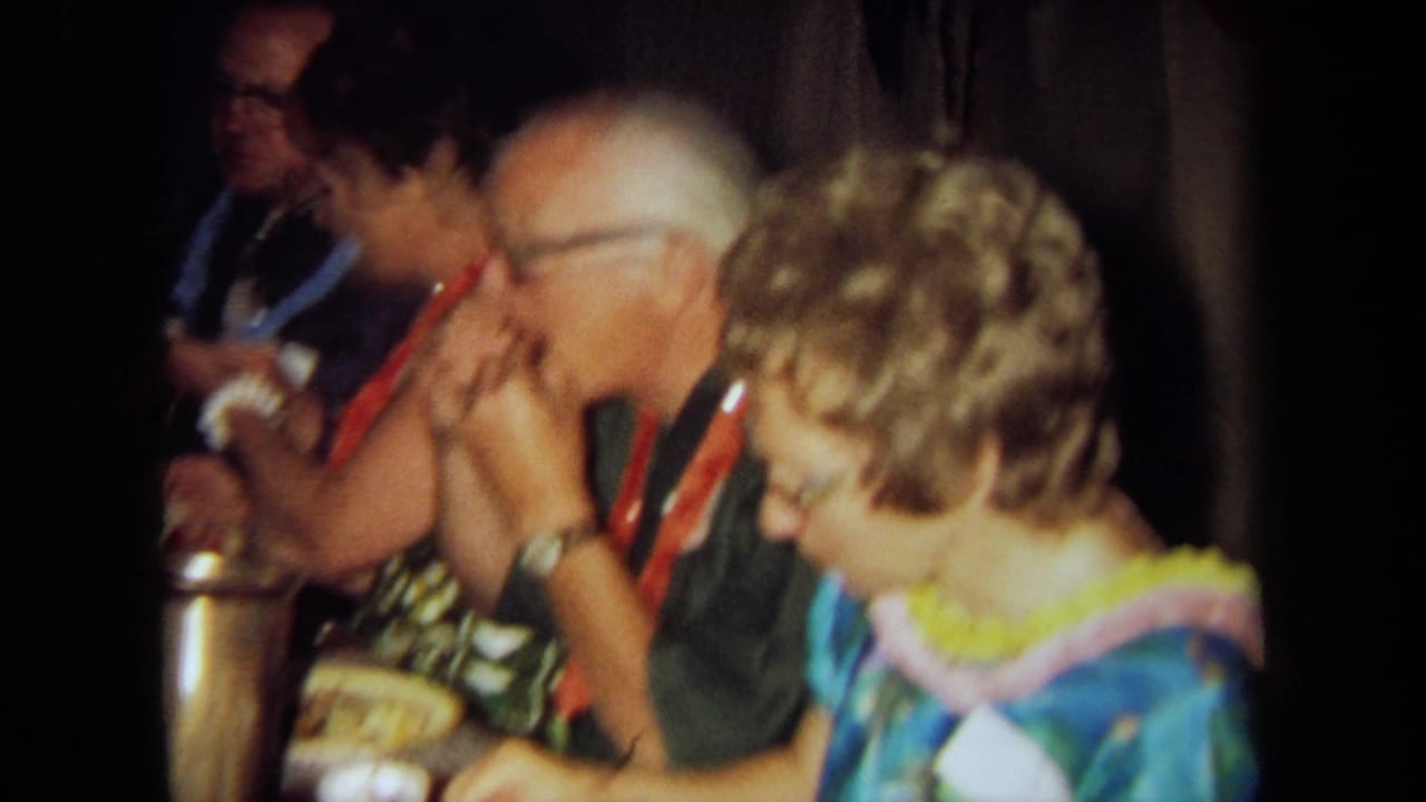 1971 California home-movie frame of adults in leis and colorful clothing seated at a crowded indoor luau-style gathering.