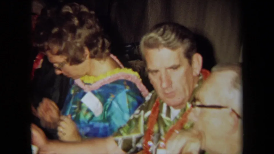 Adults wearing leis and aloha-style clothing sit at a table in a 1971 California home-movie party scene.