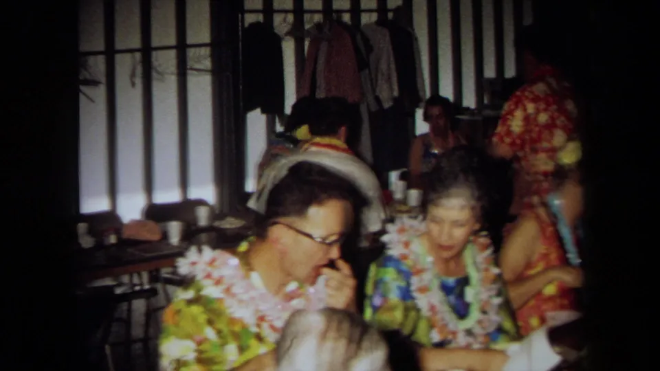 A wider 1971 California home-movie view shows adults in leis seated among tables, folding chairs, and wall panels.