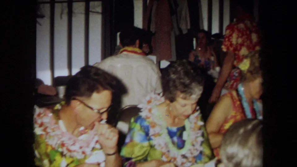 1971 California home-movie frame of luau guests in leis around tables, with folding chairs and paneled walls behind them.