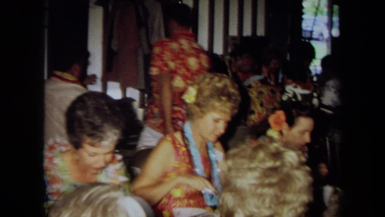 1971 California home-movie frame of adults in Hawaiian-style clothing and leis seated closely at indoor tables.