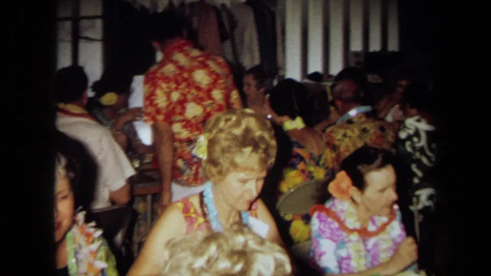 Adults wearing leis and bright Hawaiian-style clothing sit and converse at tables in a private California interior.