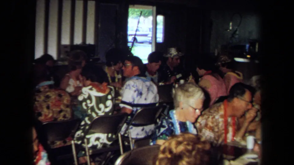 Home-movie view of adults seated at folding chairs and tables, eating and talking in a dim 1971 California gathering.