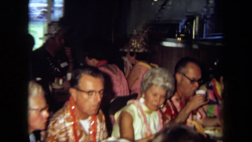 Close home-movie frame of adults in leis and patterned clothing seated at a table inside a private California room.