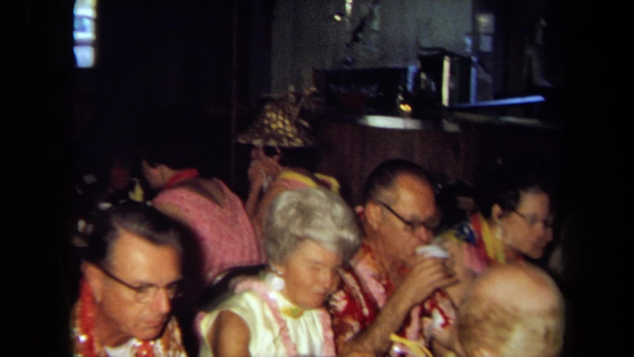 1971 California 8mm home-movie frame of adults in leis seated at a crowded indoor party table, with drinks and food visible.