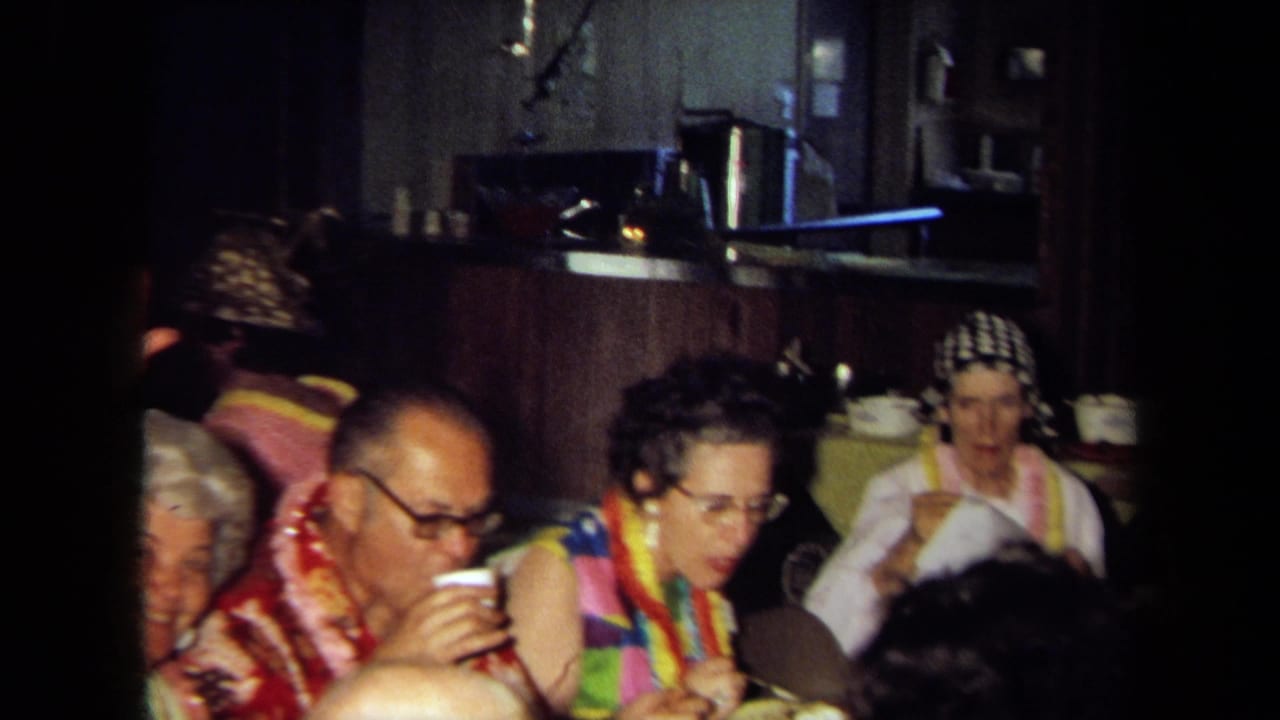 Party guests in leis sit at a table while one man drinks from a cup.