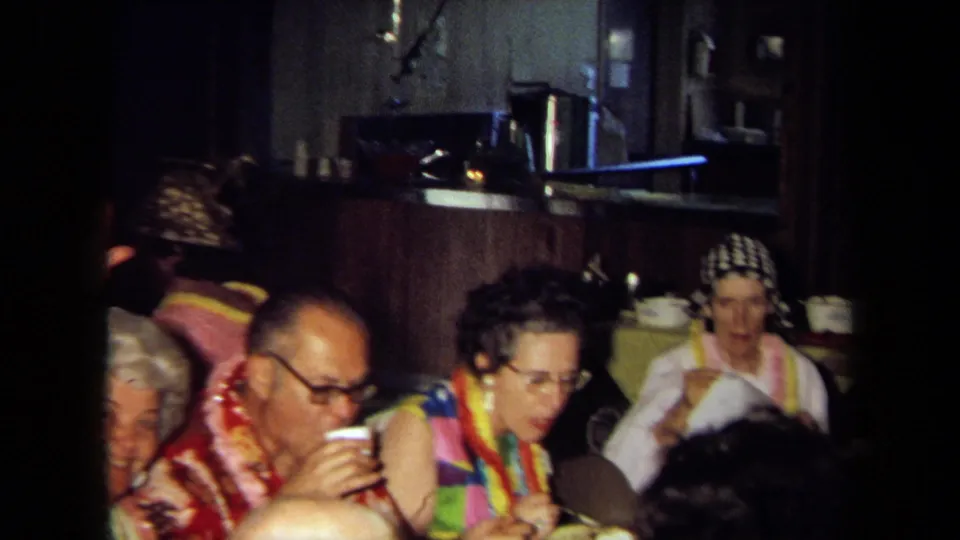 1971 California home-movie frame showing adults in leis at an indoor party table, including a man drinking from a cup.