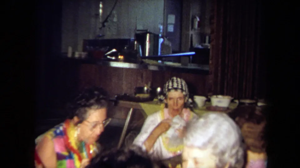 1971 California 8mm frame of seated adults at a Hawaiian-themed indoor party, with tableware, serving dishes, and leis visible.