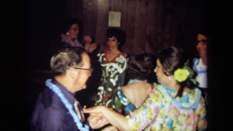 1971 California home-movie frame showing adults standing and seated in conversation at a Hawaiian-themed indoor party.