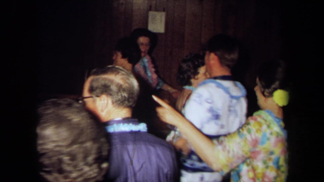 1971 California 8mm frame of older adults in leis and colorful clothing gathered in a dim private party room.