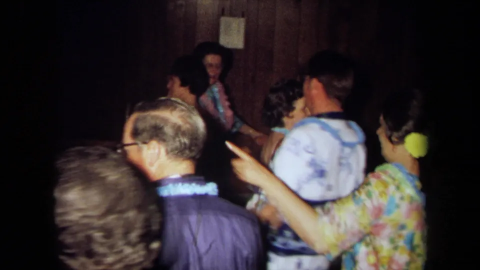 1971 California 8mm frame of older adults in leis and colorful clothing gathered in a dim private party room.