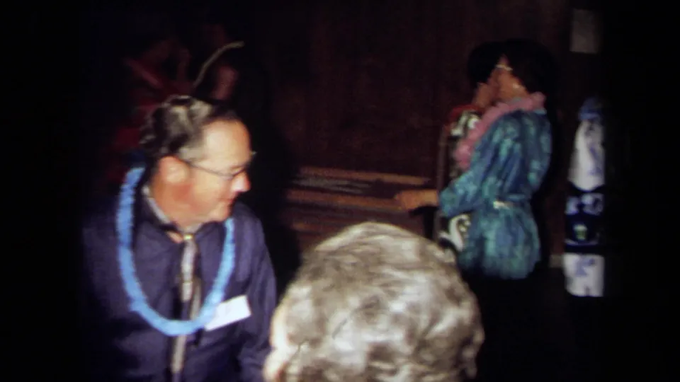 1971 California party frame showing an older man in glasses, a purple shirt, and blue lei among guests in a dark interior.