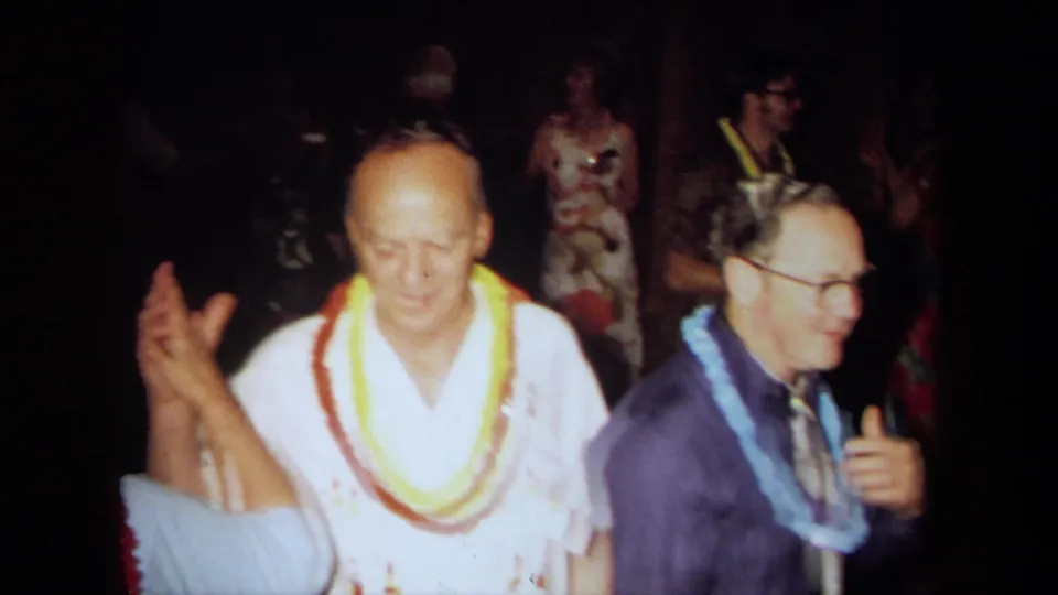 1971 California 8mm frame of older men wearing colorful leis dancing at a Hawaiian-themed indoor party.