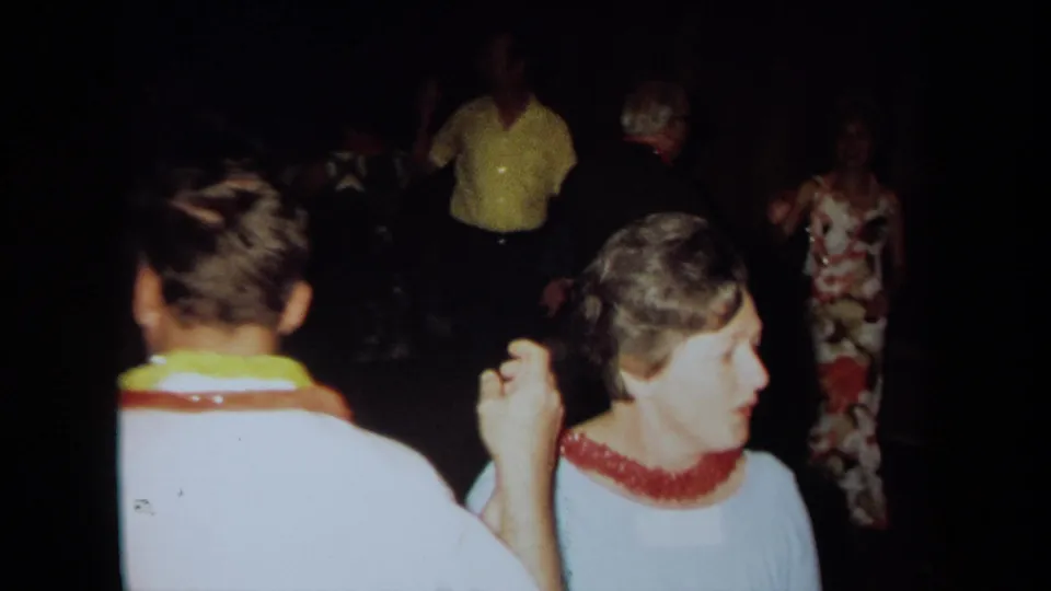 1971 California home-movie frame of lei-wearing guests dancing in a dim private interior during a themed party.