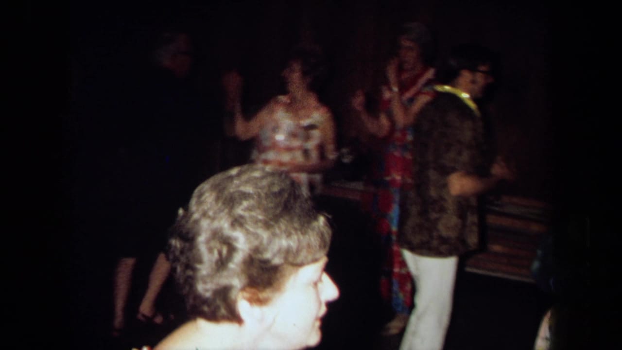 1971 California home-movie frame showing adults dancing and socializing in a dim private interior with wood-paneled walls.
