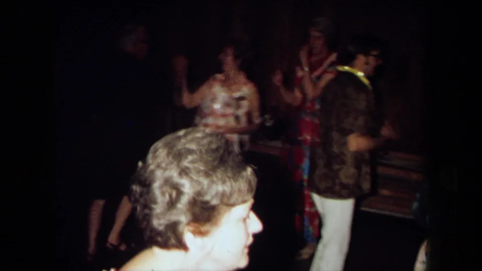 1971 California home-movie frame showing adults dancing and socializing in a dim private interior with wood-paneled walls.