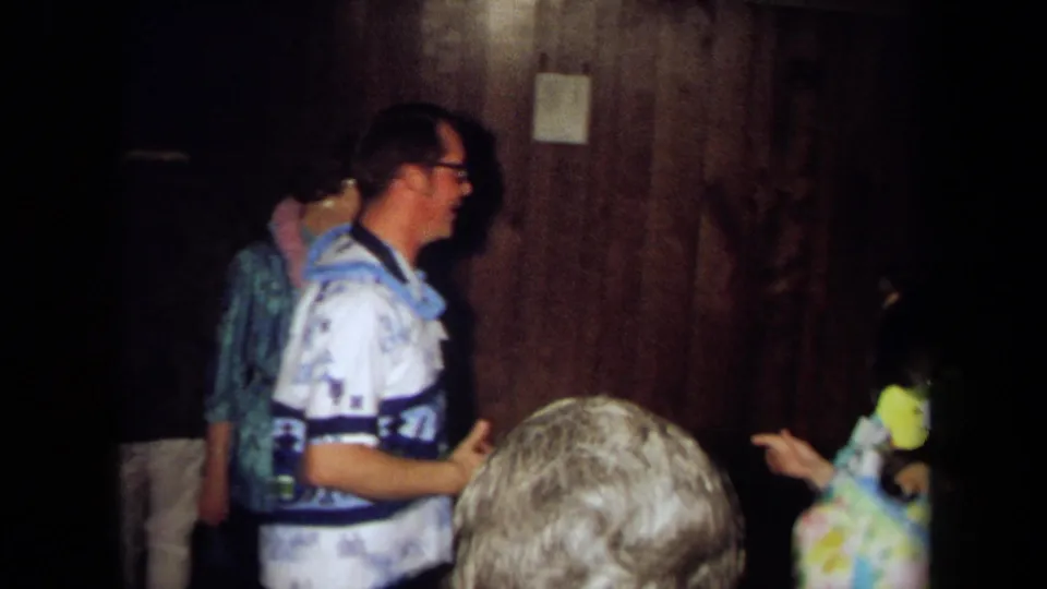 A man in a patterned blue and white shirt smiles and gestures among adults in a 1971 California indoor party scene.