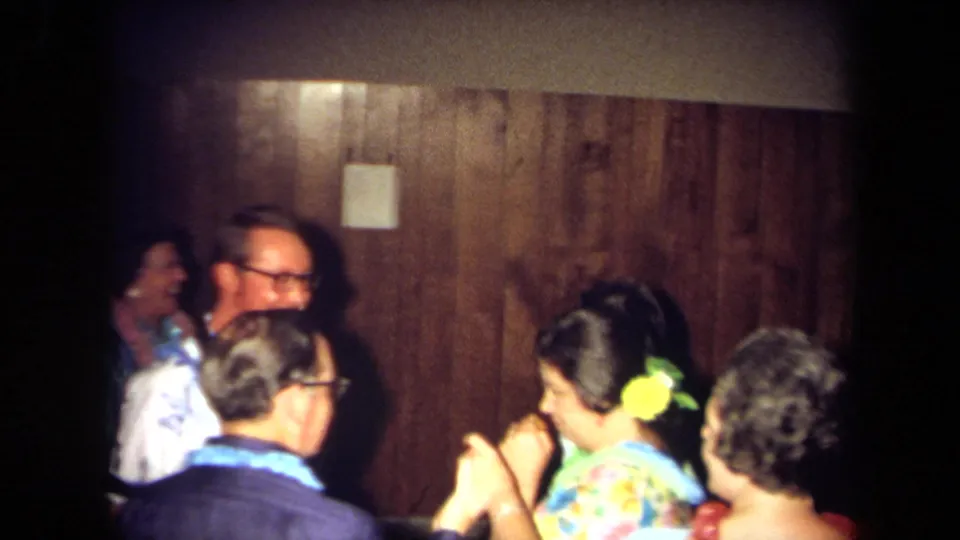 Adults in 1970s clothing converse and move together in a dim California home interior with wood paneling.