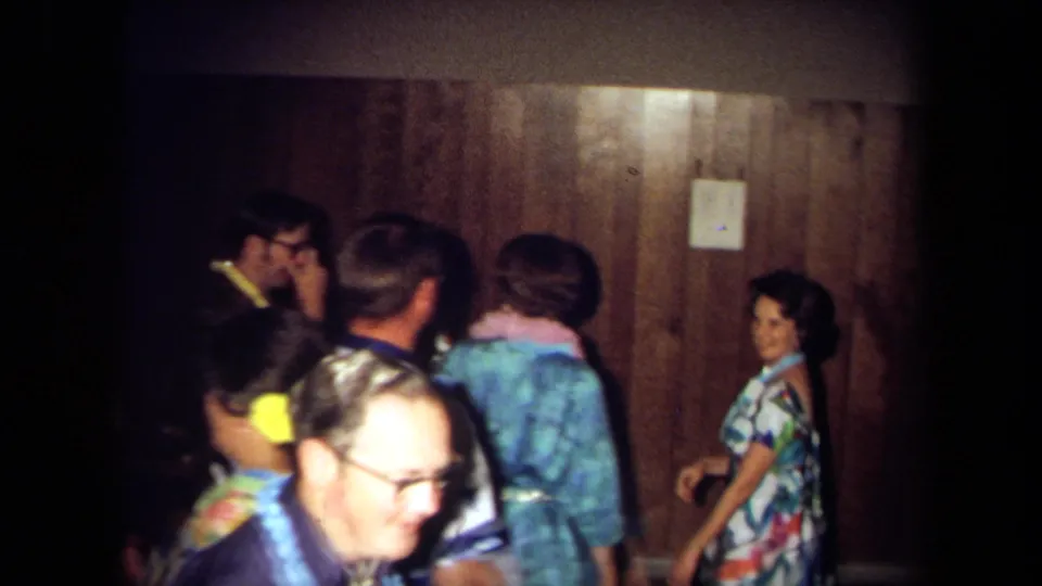 1971 California home-movie frame of adults dancing and socializing near wood paneling, with patterned clothing and visible faces.