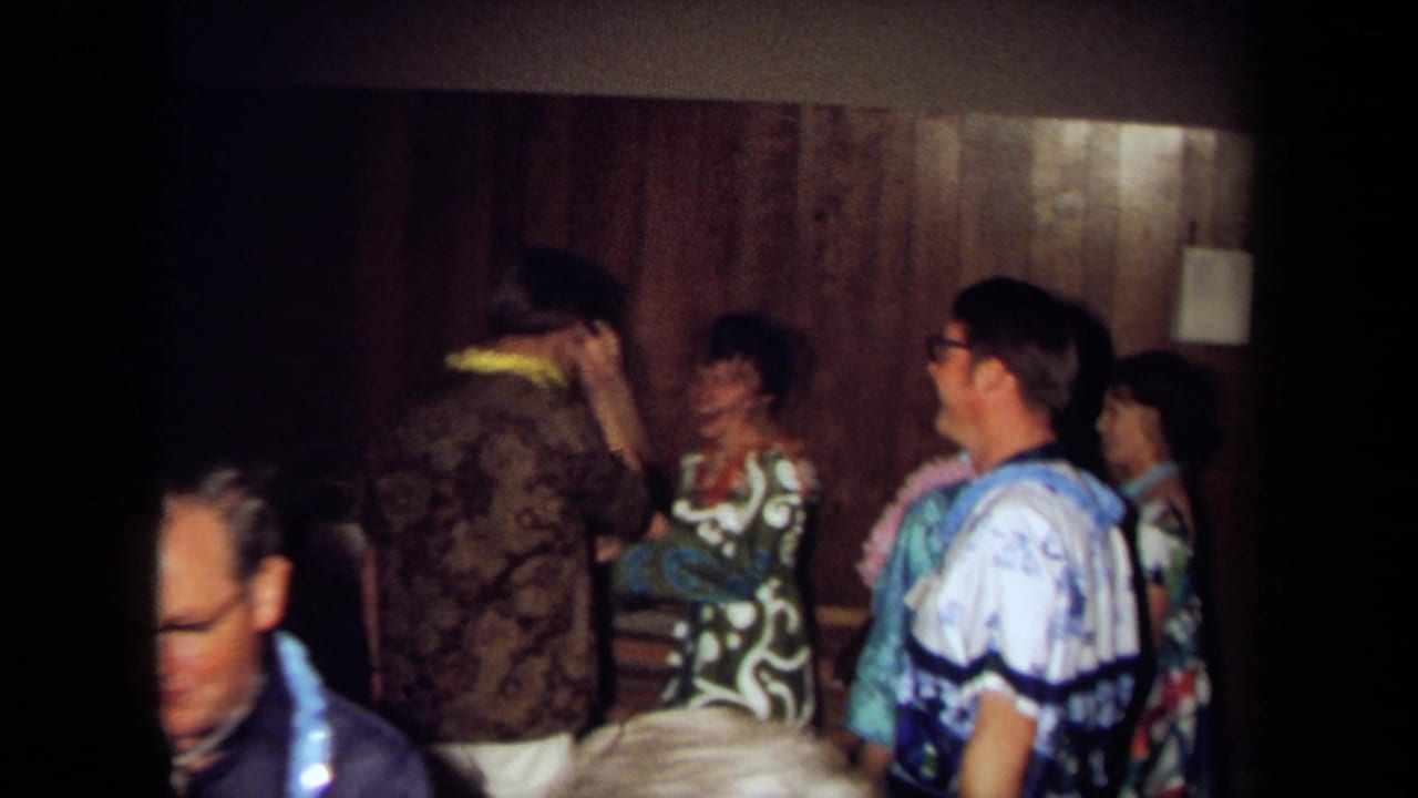 1971 California home-movie frame showing adults in leis and patterned clothing gathered in a dim wood-paneled party room.