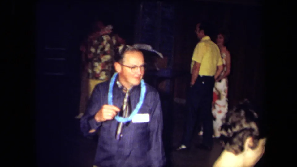 A man in glasses and a blue lei stands in a dim 1971 California party setting while other adults mingle behind him.