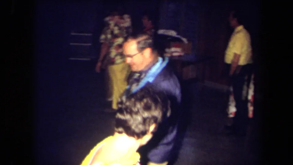 Handheld 1971 California frame of a man in a blue lei moving near a woman in a yellow lei with party guests in the background.