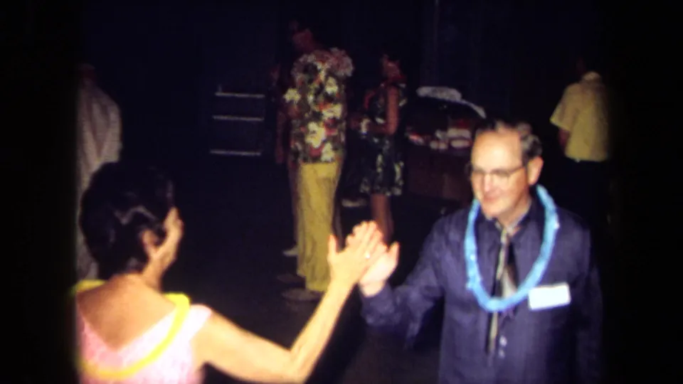 A woman in a pink dress and yellow lei dances with a smiling man in glasses and a blue lei at a 1971 California gathering.