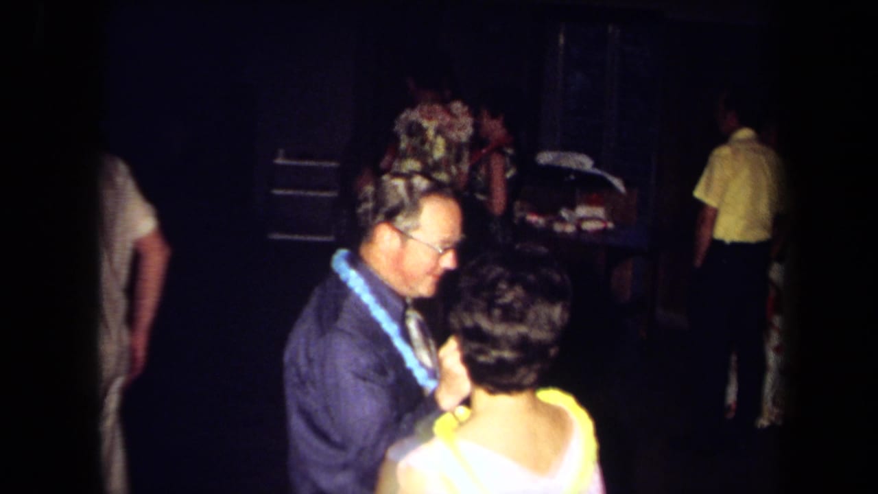 A man with a lei dances with a woman in a light dress while other party guests stand in the background.