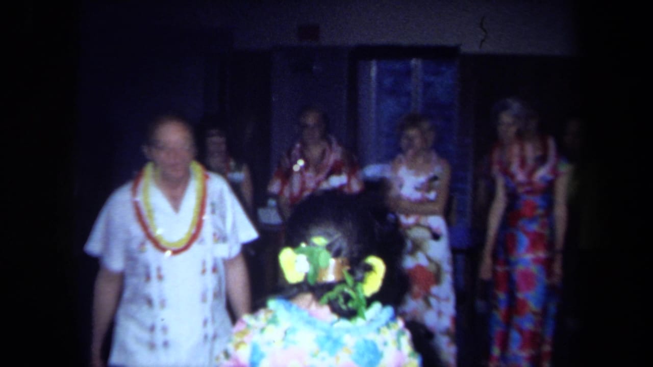 1971 California home-movie frame showing adults in Hawaiian-themed attire, with leis and floral dresses in a dim indoor room.
