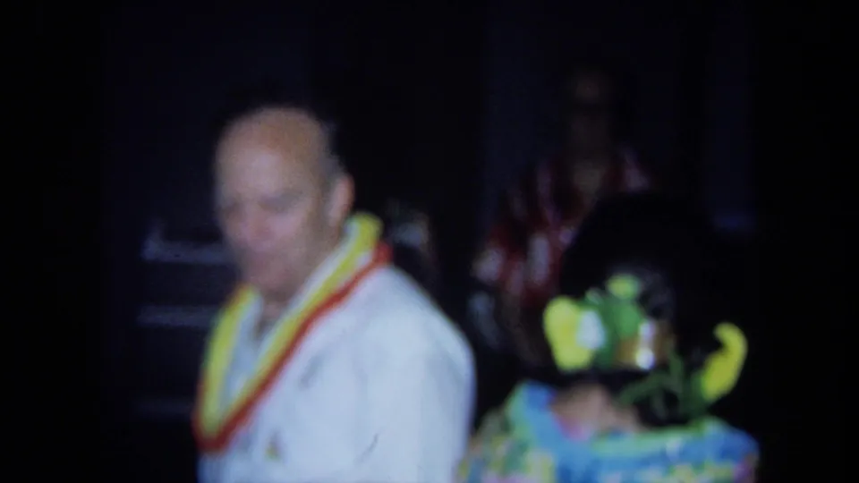 Soft 8mm frame from the 1971 California party showing a man in a white shirt and lei beside a brightly dressed dancer.