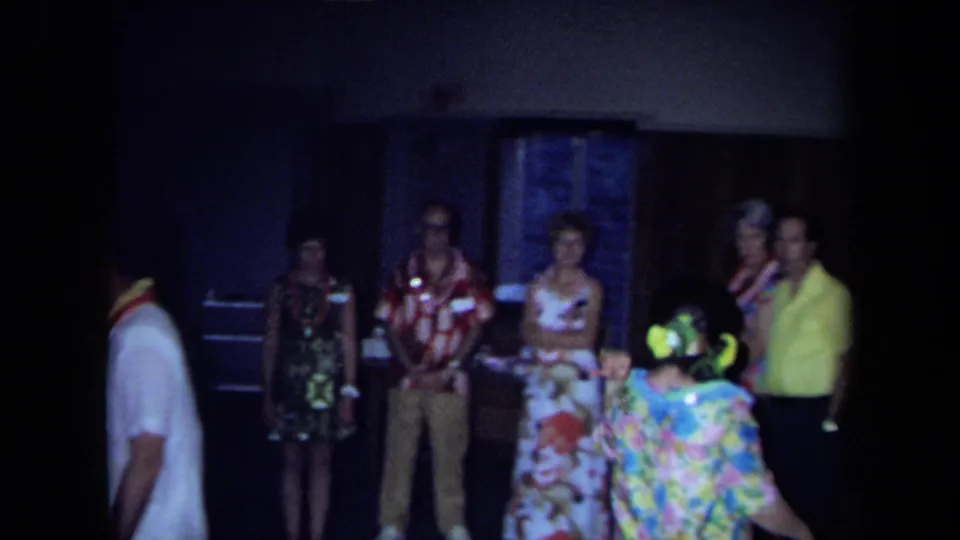 1971 California home-movie frame with adults in leis and patterned clothing watching a dancer in a dim wood-paneled room.