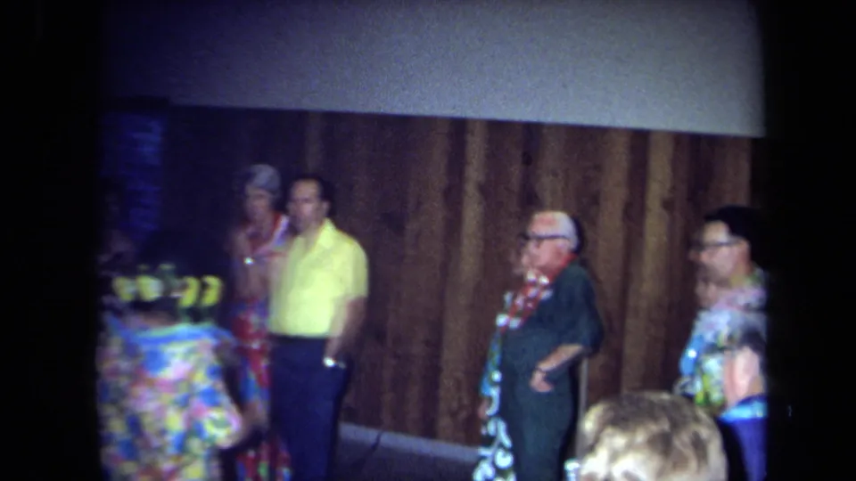 1971 California party frame showing adults in leis, colorful clothing, and glasses near wood paneling during a Hawaiian-themed gathering.