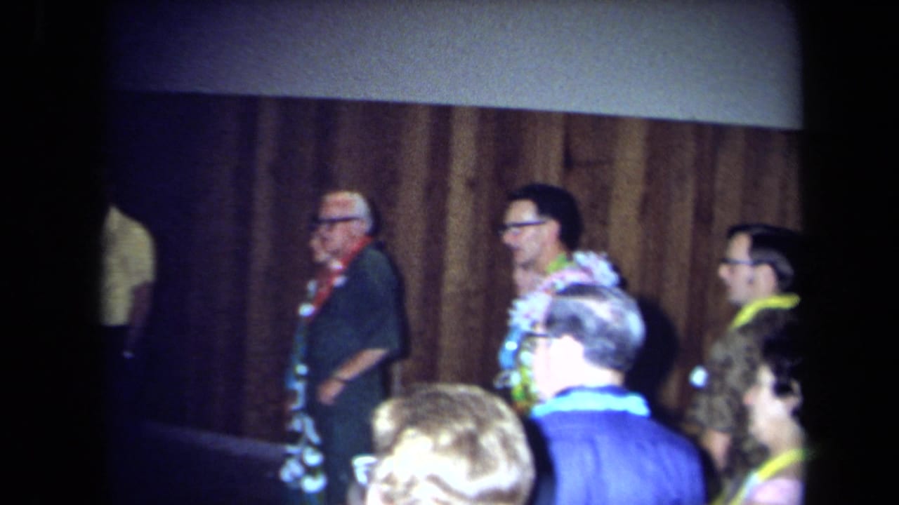 1971 California home-movie frame showing adult guests in leis and patterned clothing near wood paneling during an indoor party.