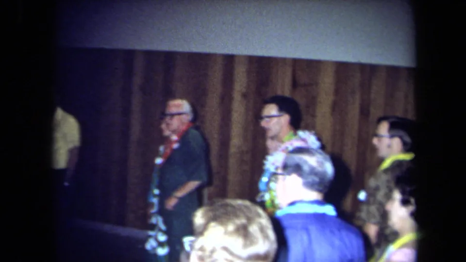 1971 California home-movie frame showing adult guests in leis and patterned clothing near wood paneling during an indoor party.