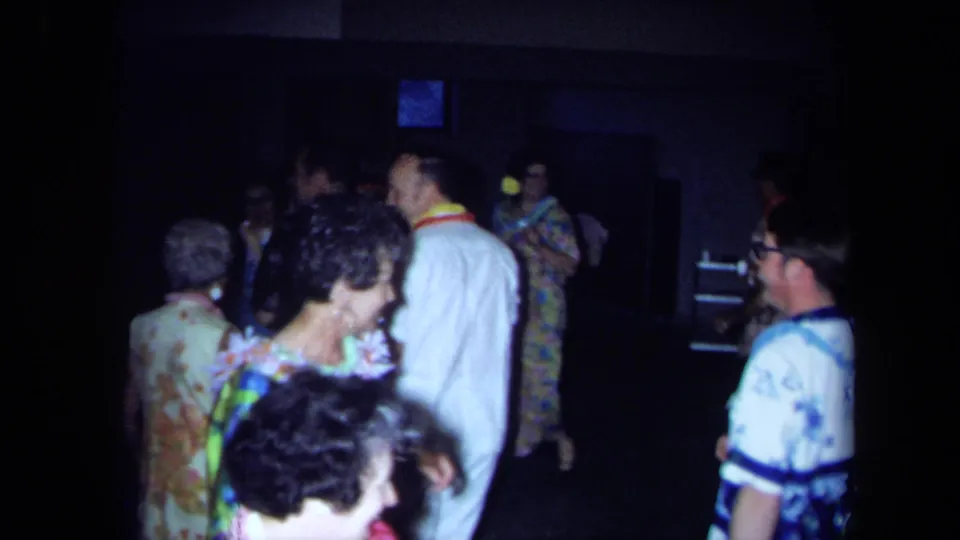 1971 California frame showing adult party guests in leis, aloha-style shirts, and floral clothing on a dim dance floor.