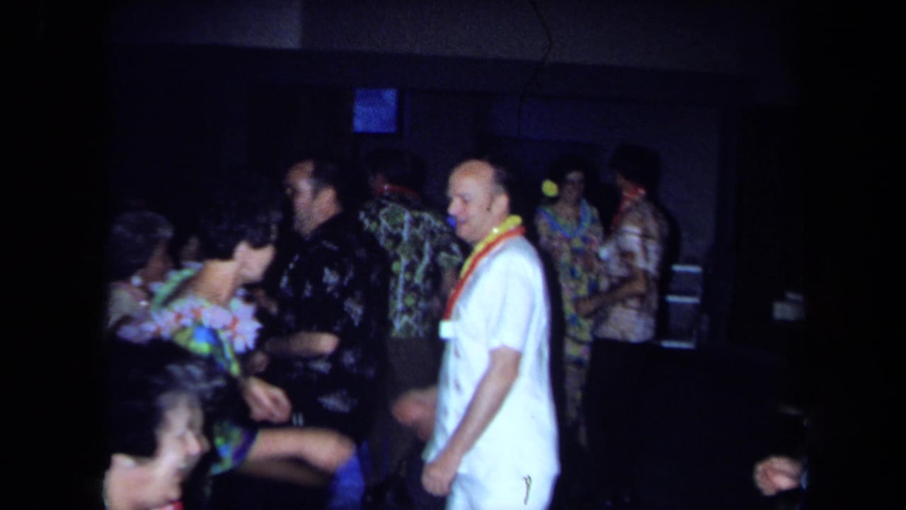 1971 California home-movie frame showing adults in leis, aloha shirts, and floral clothing dancing inside a dim private room.