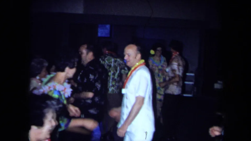 1971 California home-movie frame showing adults in leis, aloha shirts, and floral clothing dancing inside a dim private room.