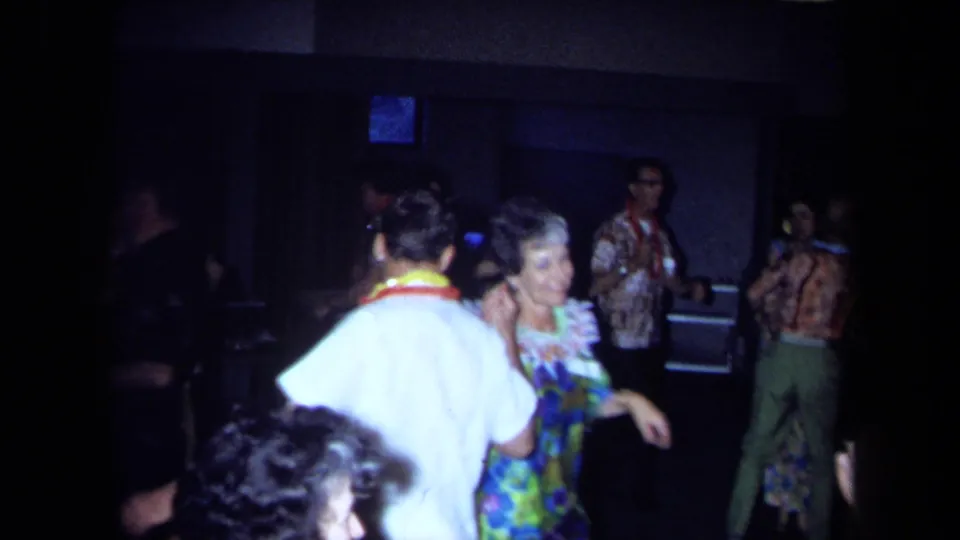 Adults at a 1971 California Hawaiian-themed party dance and talk, with leis and patterned shirts visible in the dark room.