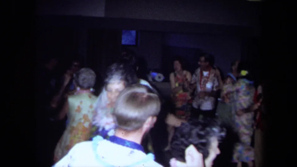 1971 California home-movie frame of a crowded indoor party with adults in leis and floral clothing, plus a child seen from behind.