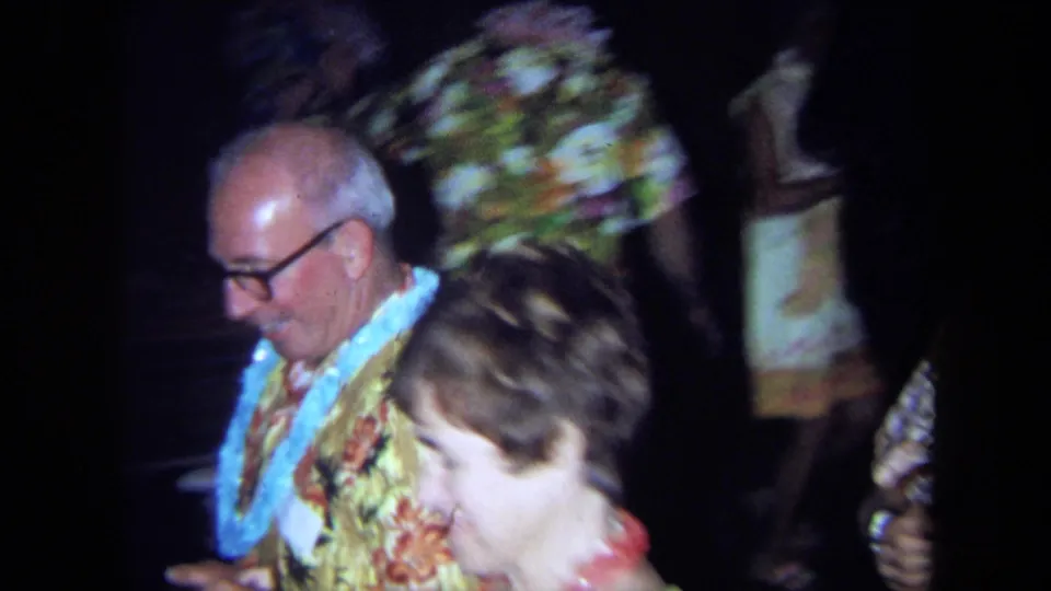 Close handheld 1971 California party frame showing a man with glasses and a lei dancing near other guests in patterned clothing.