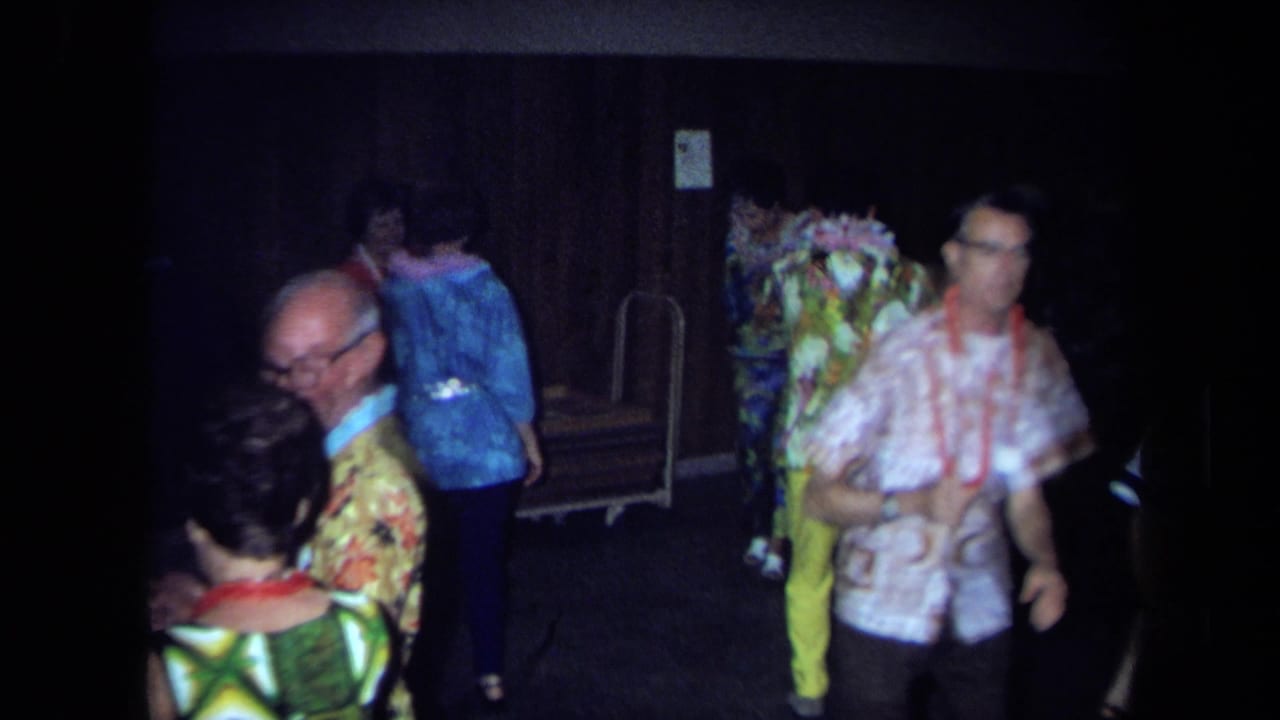 1971 California home-movie frame showing adults in leis and patterned clothing dancing and mingling indoors.