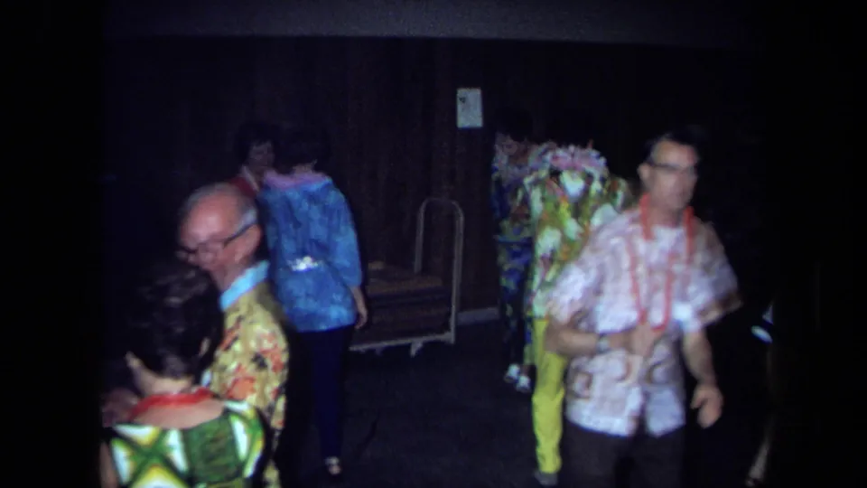 1971 California home-movie frame showing adults in leis and patterned clothing dancing and mingling indoors.