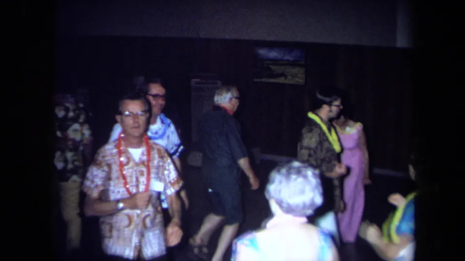 Adults at a 1971 California indoor party stand and move through the frame, several wearing leis and bright patterned outfits.