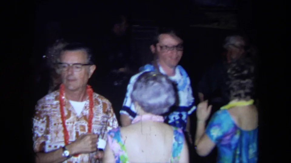 1971 California party frame with adults in leis, eyeglasses, and colorful clothes gathered close to the handheld camera.