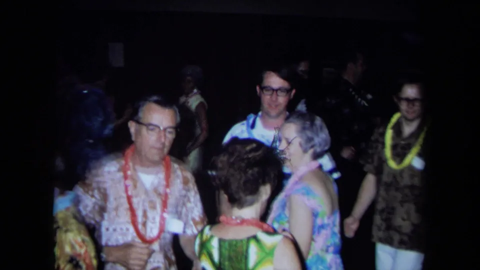 Adults at a 1971 California indoor gathering converse and dance, with leis, floral clothing, and soft 8mm exposure visible.