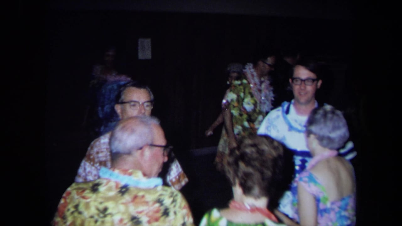 1971 California home-movie frame showing adults in leis and bright patterned clothes dancing in a dark interior.