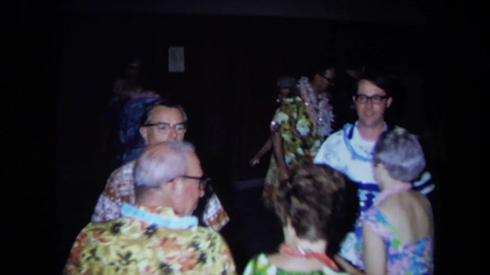 1971 California home-movie frame showing adults in leis and bright patterned clothes dancing in a dark interior.